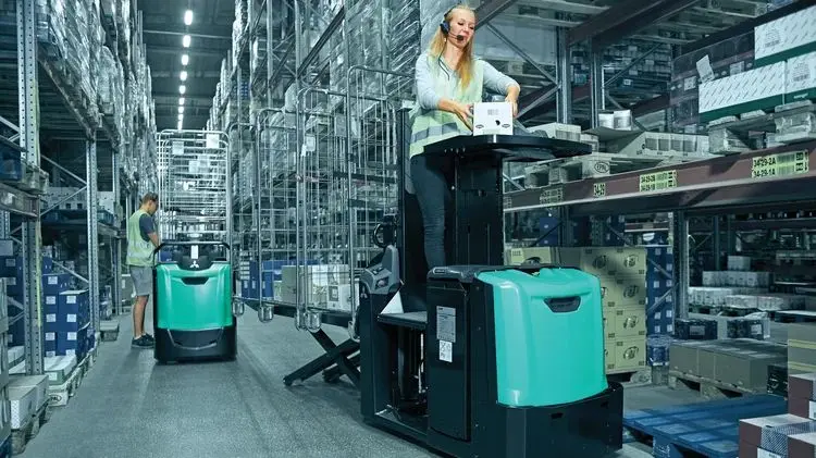 Lagermedarbetare med headset som plockar gods från en hylla stående på en Logisnext låglyftande orderplocktruck i en lagergång